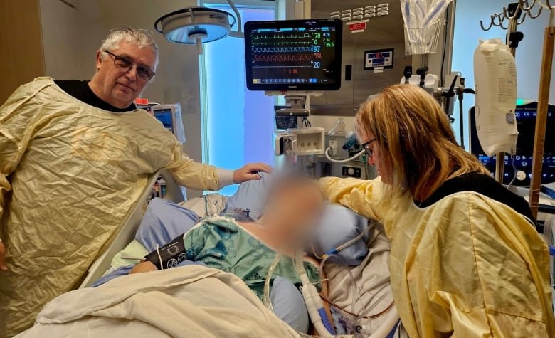 An older man and woman stand next to a hospital bed where a figure is surrounded by tubes and equipment. The patient's face is blurred to protect their privacy.