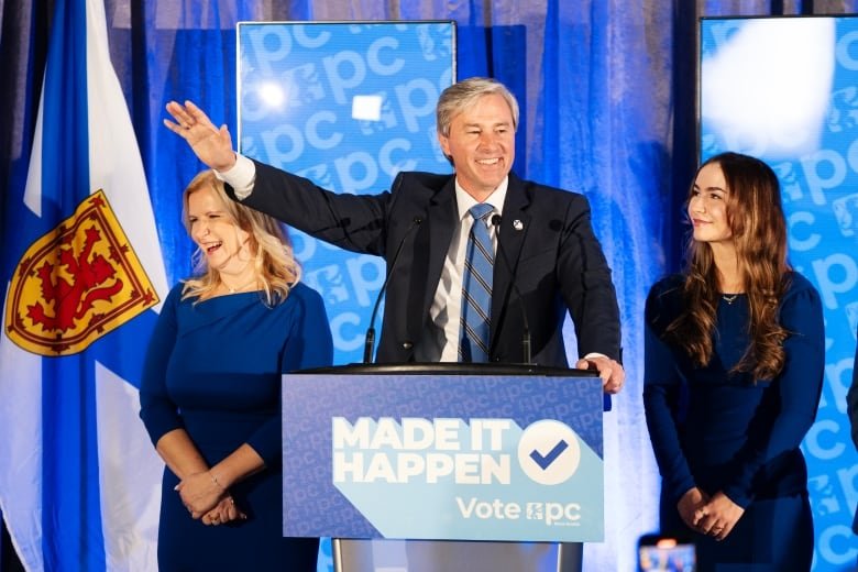 A man stands behind a podium with hand extended.