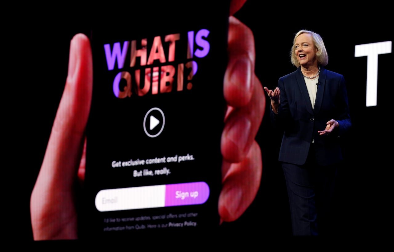 A woman in a dark pantsuit stands presenting to an audience off camera. behind her, a big screen displays a phone in hand, and hte display on the phone reads "what is quibi?"