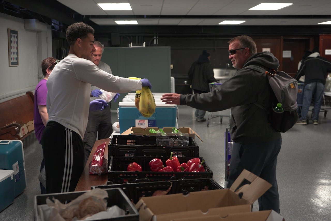 A young man passes a bag to an elderly man wearing shades and a coat, suggesting a transaction. A table with fruits and vegetables separates them.