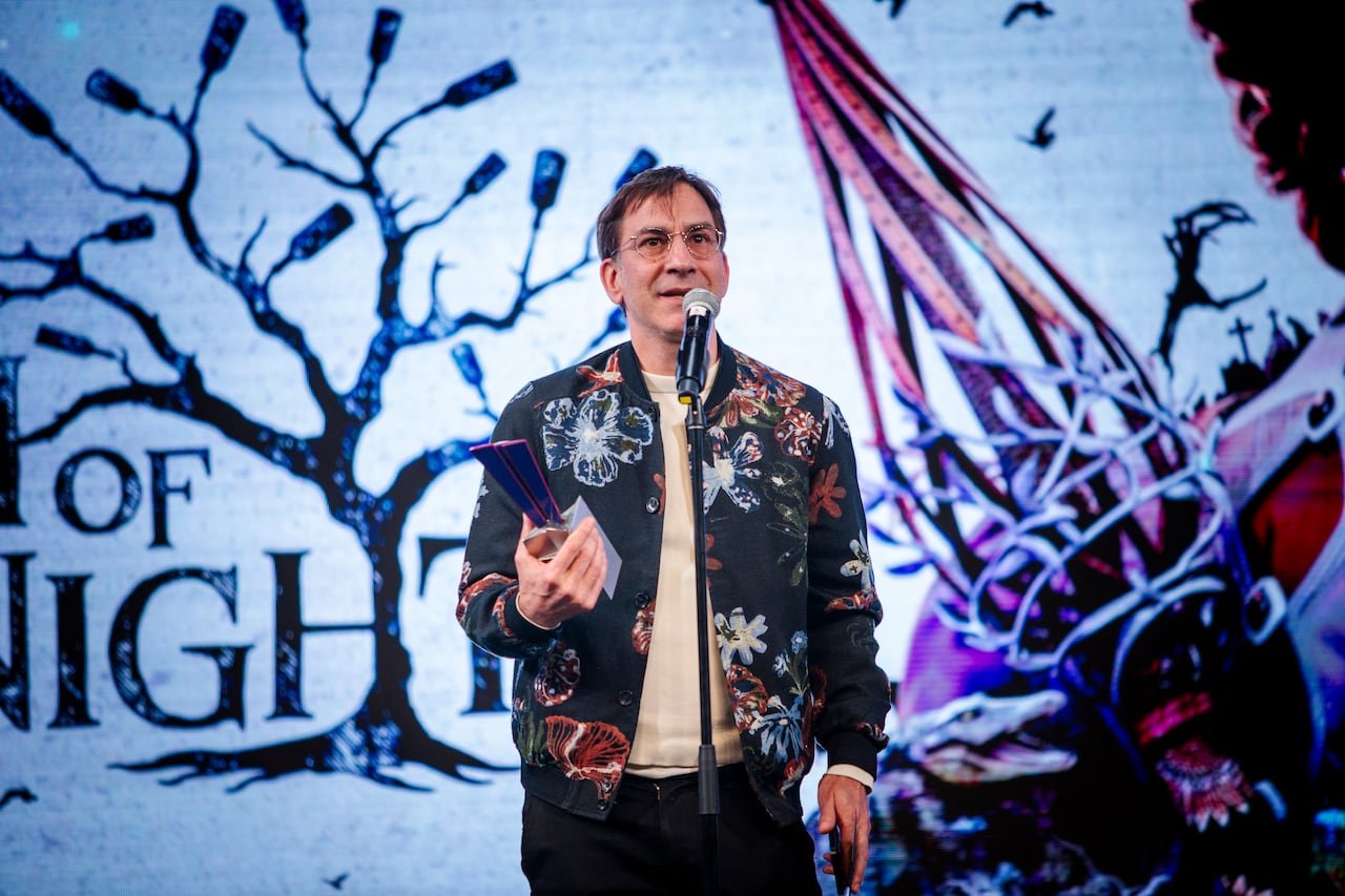 A man in a flowery jacket accepting an award on stage with a microphone in hand.
