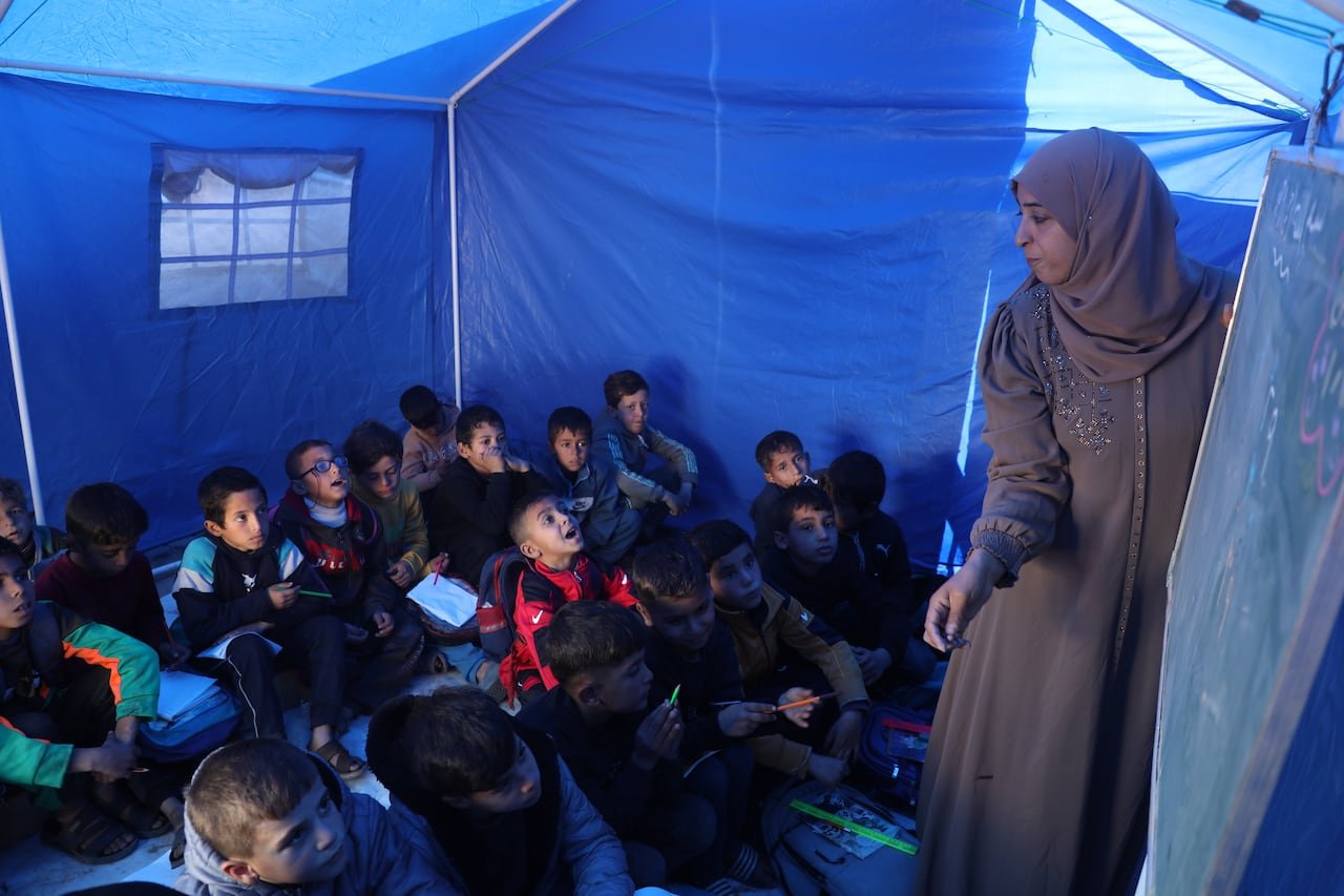 A woman in a beige attire and matching hijab writes on a chalkboard while surrounded by small children inside a blue tent.
