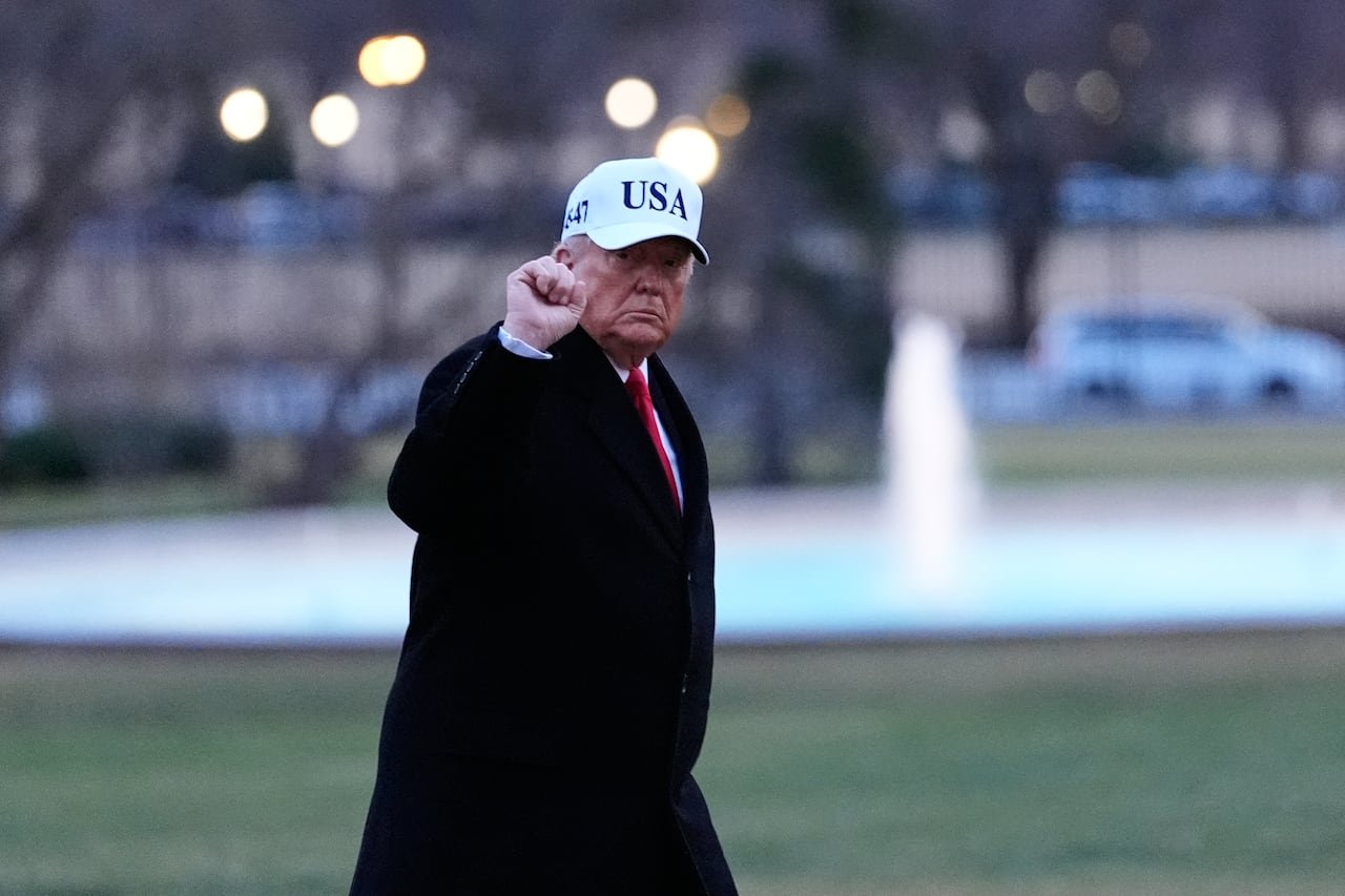 A man in a long winter overcoat and a baseball cap raises a fist while walking on a lawn.