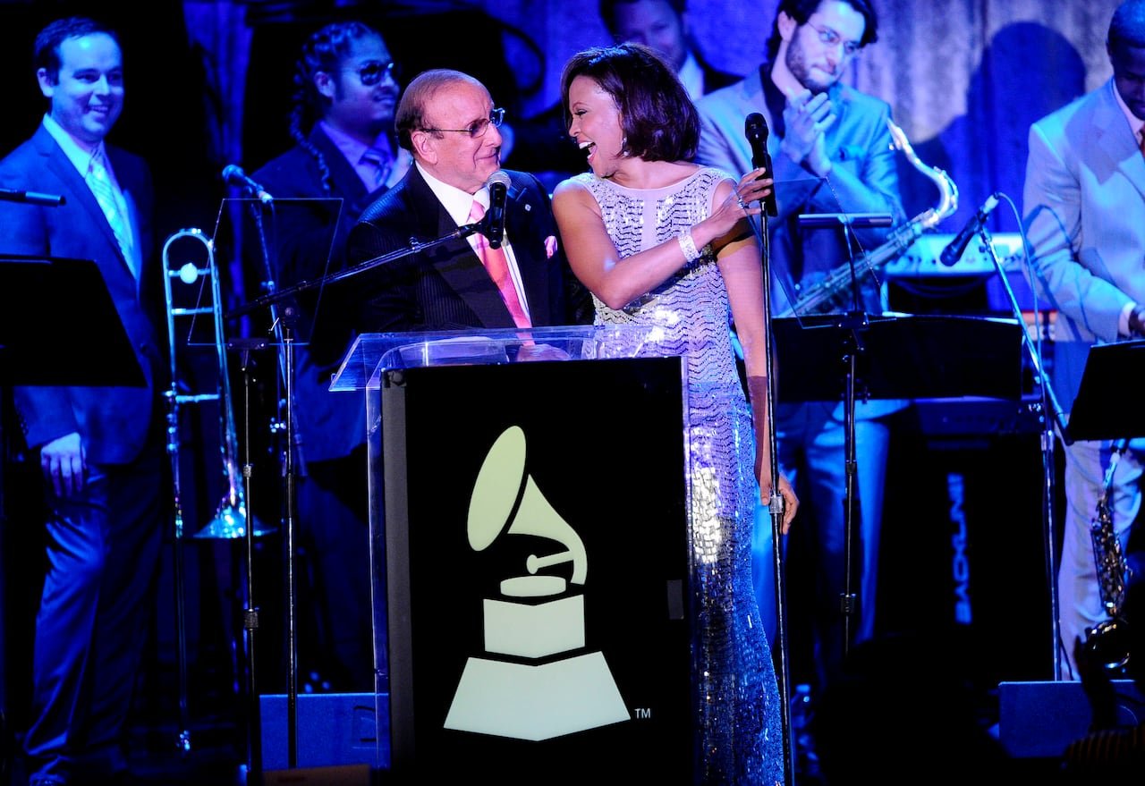 default-575 An older, bald man, wearing glasses, a black suit, white shirt and pink tie, stands next to a Black woman with her hair in a bob cut and wearing a silver gown. The two are smiling at each other on a stage, with musicians in the background.