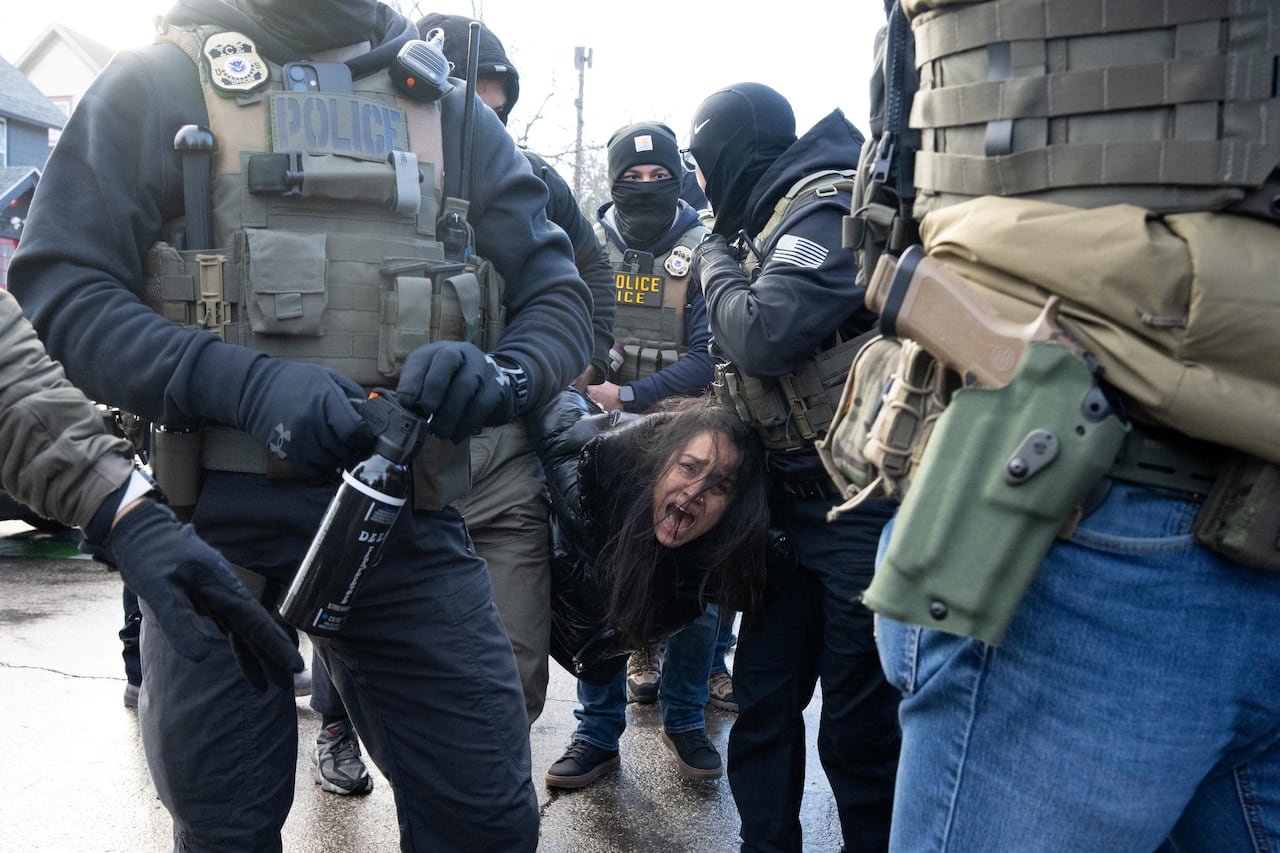 default-84 Several masked people wearing police vests hold a woman in a parka, her long, dark hair partially covering her face as she yells.
