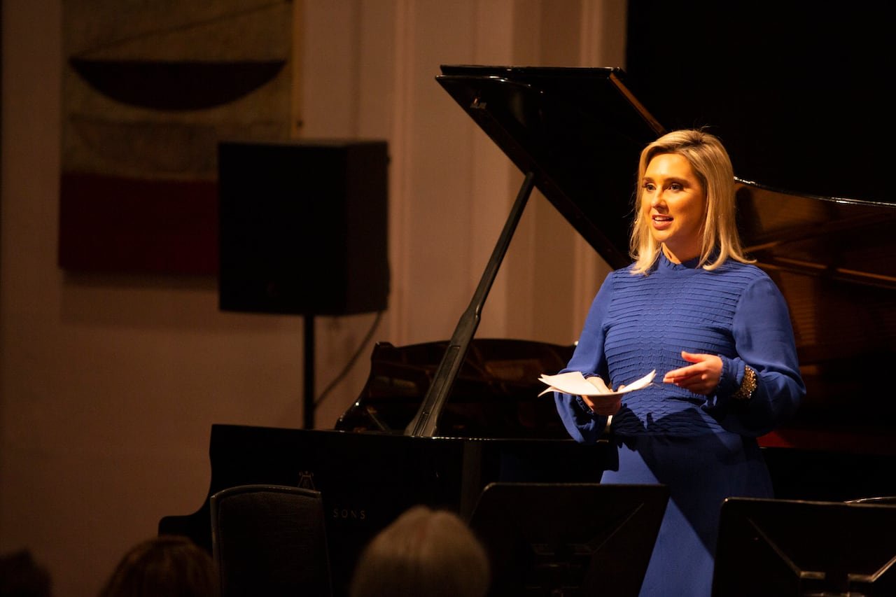 A woman addresses an audience with a grand piano in the background.